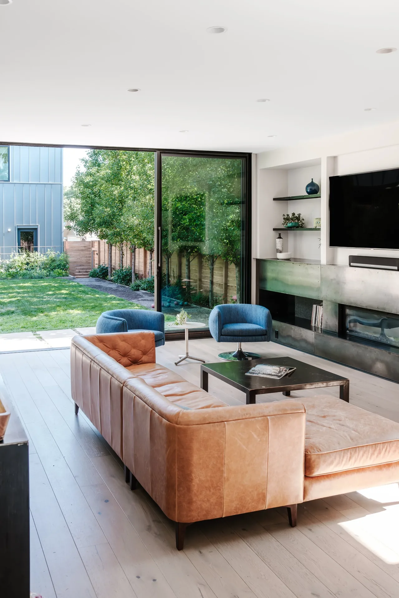 Modern living room features a tan leather sectional, blue chairs, and a wall-mounted TV beside sliding glass doors.