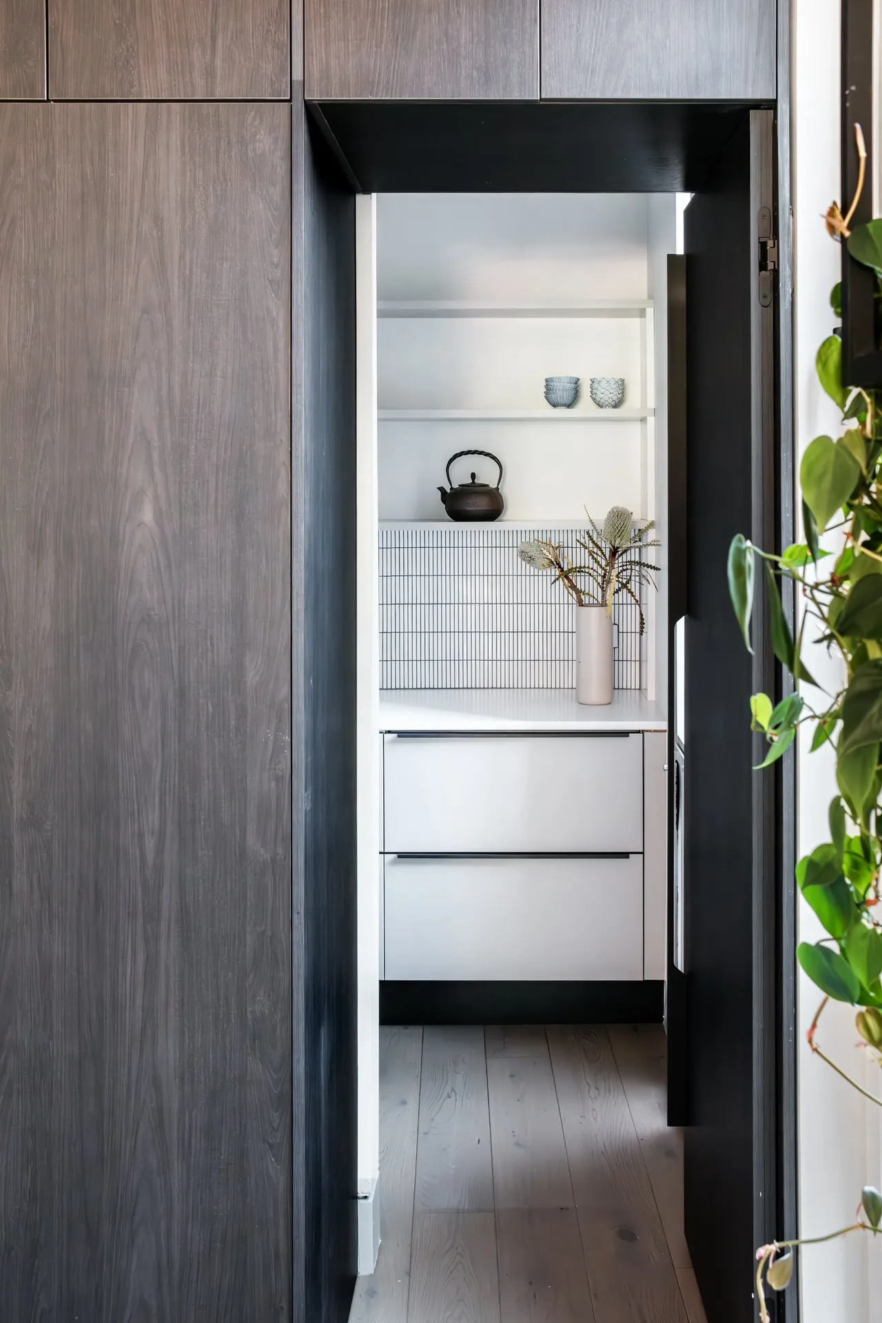 White drawers and open shelves with a black teapot and cups inside a kitchen nook framed by dark wood cabinetry.
