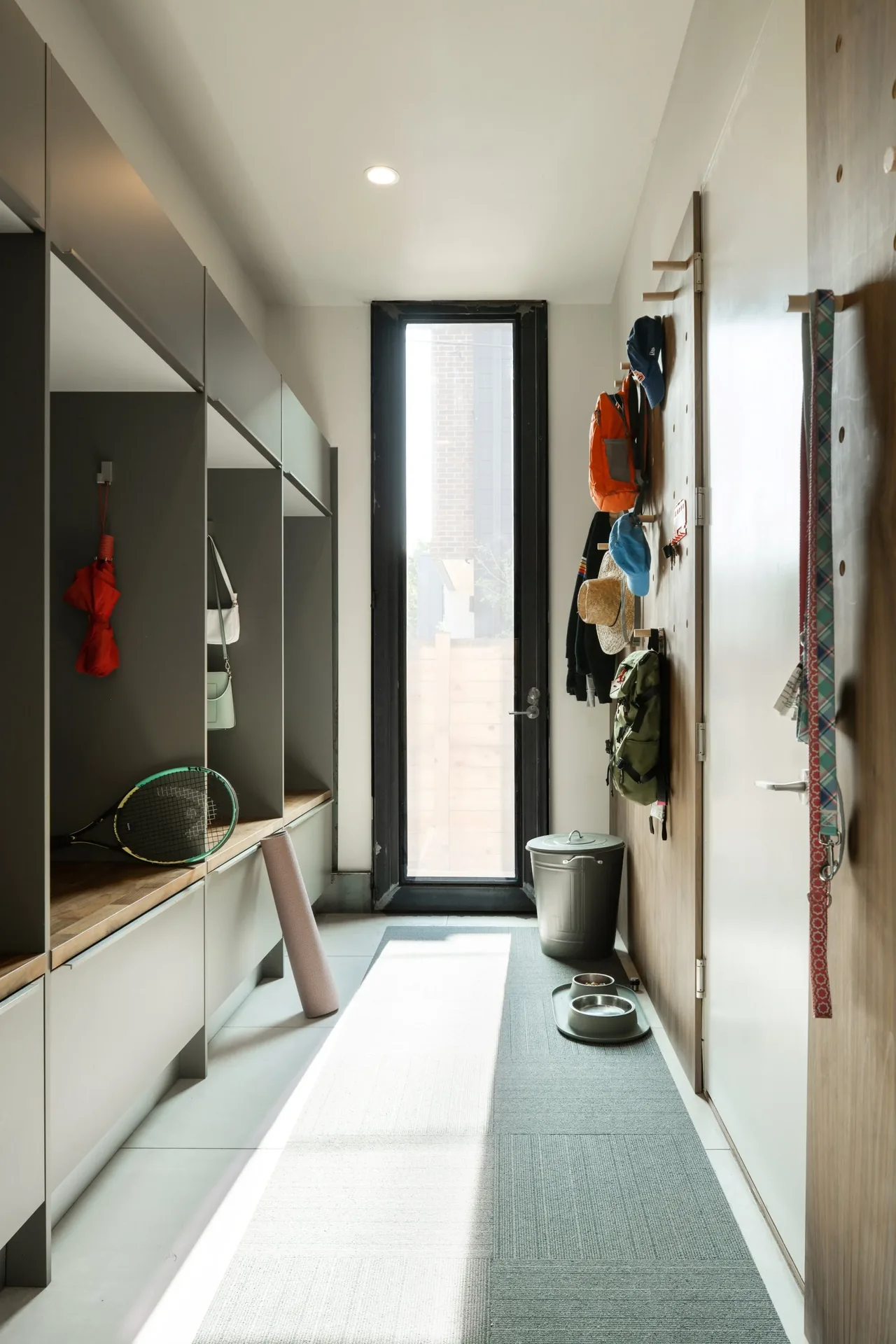 Mudroom with gray built-in storage, a tall black-framed glass door, and wall hooks with bags and hats.