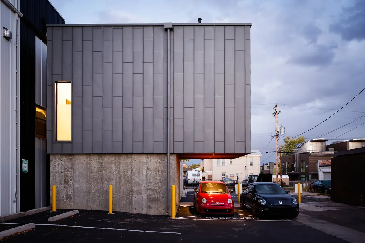 The alley house features a cantilevered upper level with metal cladding above a concrete base, including parking space beneath.