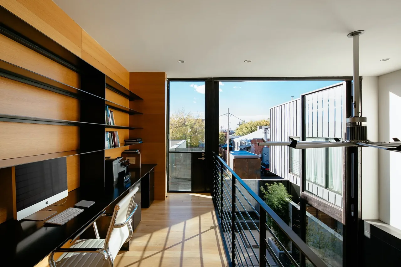 A modern home office space with built-in wood shelves, black metal railing, and a balcony door providing outdoor views.