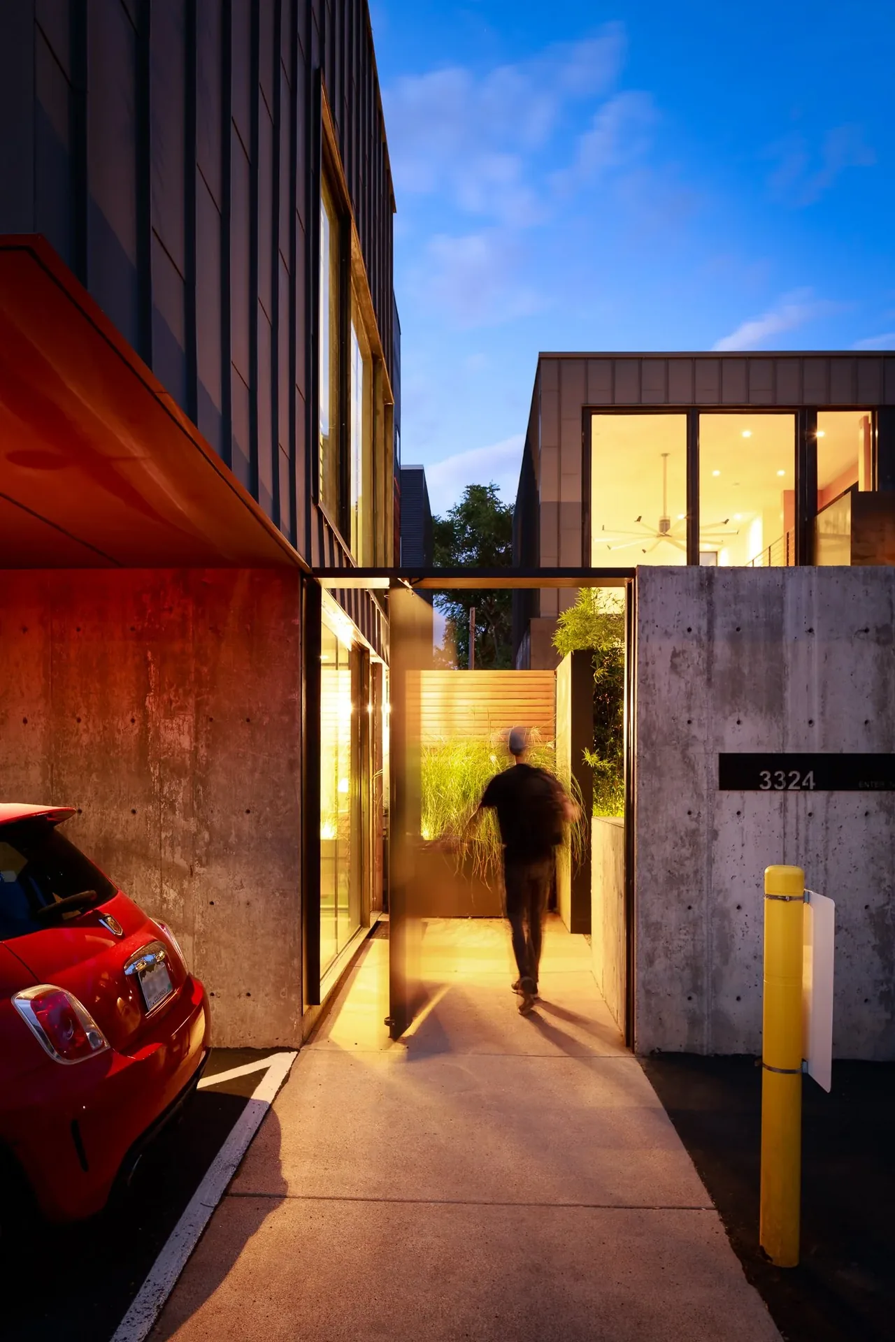 Entry gate of Alley House featuring concrete walls and large windows illuminated at dusk.