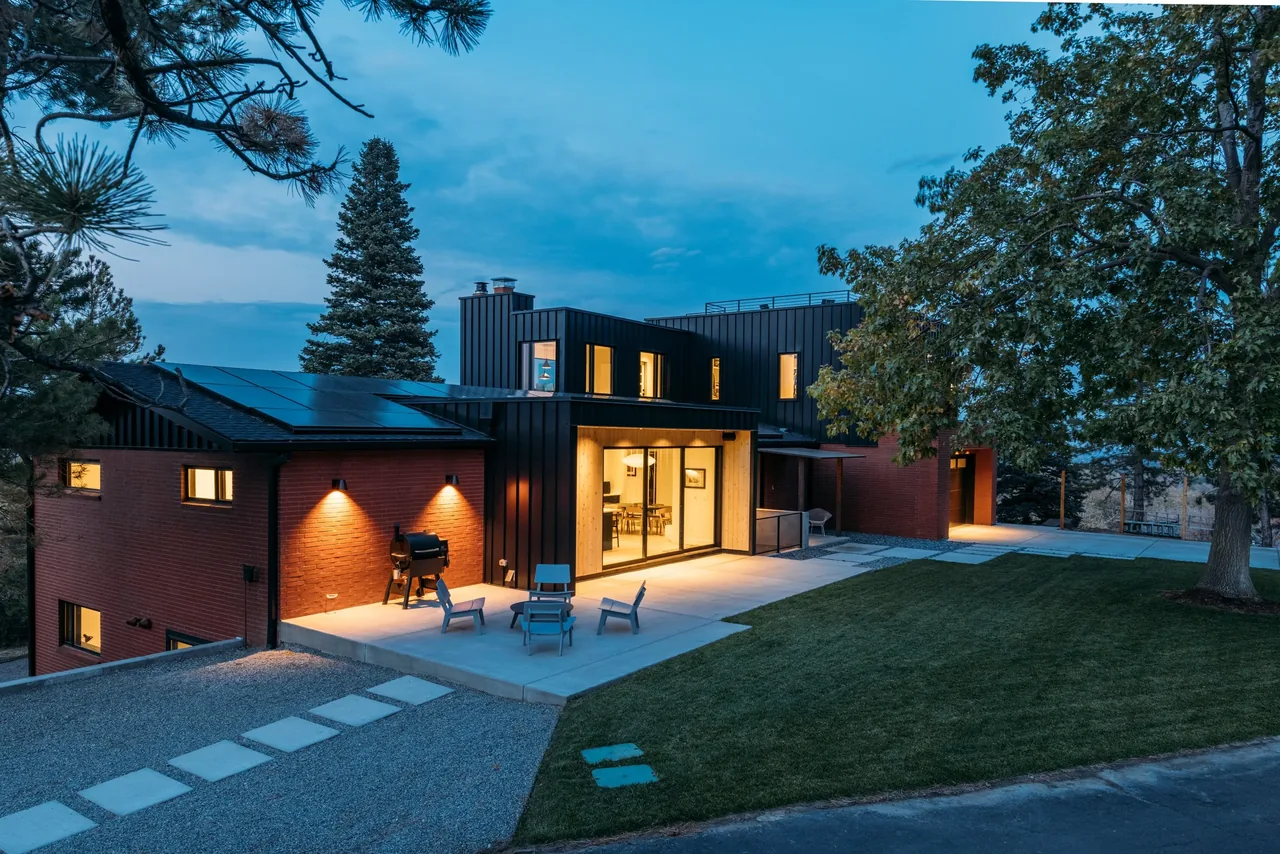 Modern two-story home with black metal and brick exterior, illuminated outdoor wall lights, and a concrete patio with seating in a forested setting.