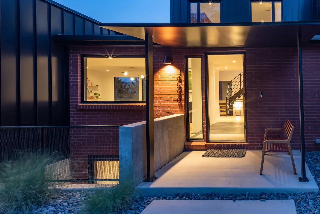 Modern front entry with brick wall, black metal siding, and warm lighting highlights the covered concrete patio and wooden bench.