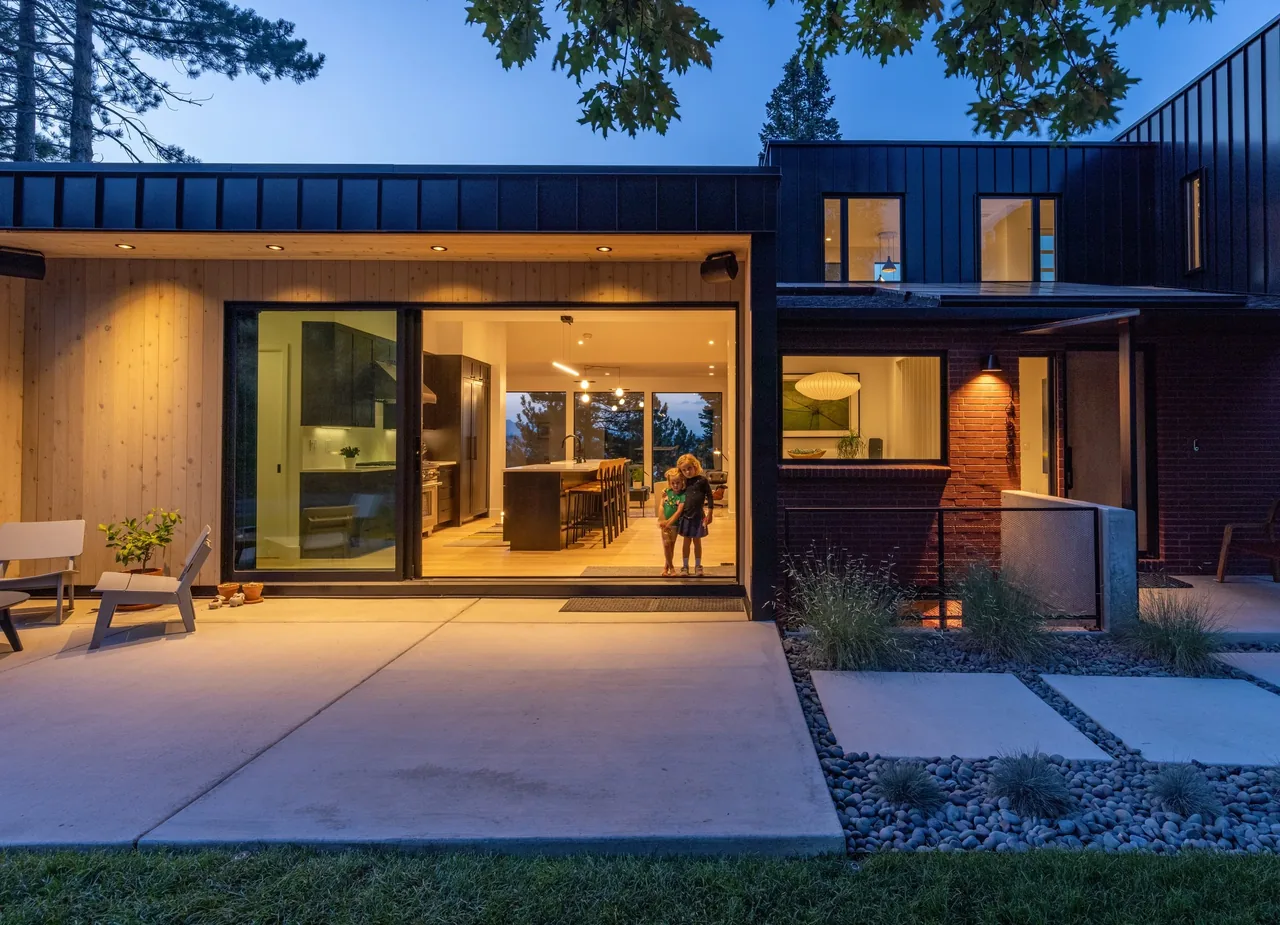 Modern home's patio with sliding glass doors revealing kitchen and dining, featuring black window frames and warm exterior lighting.