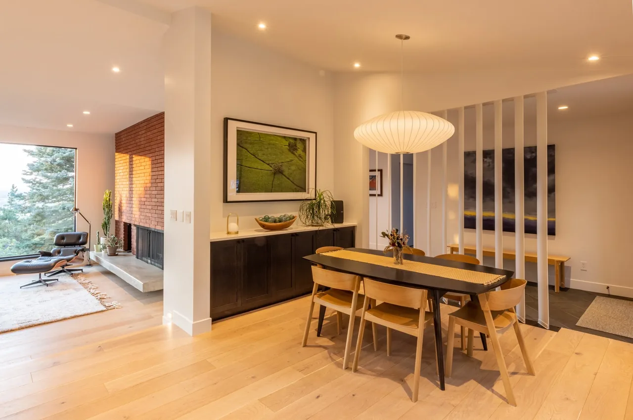 Dining room with black cabinetry, light wood floors, and a large round pendant light near a living area with a concrete hearth and leather lounge chair.