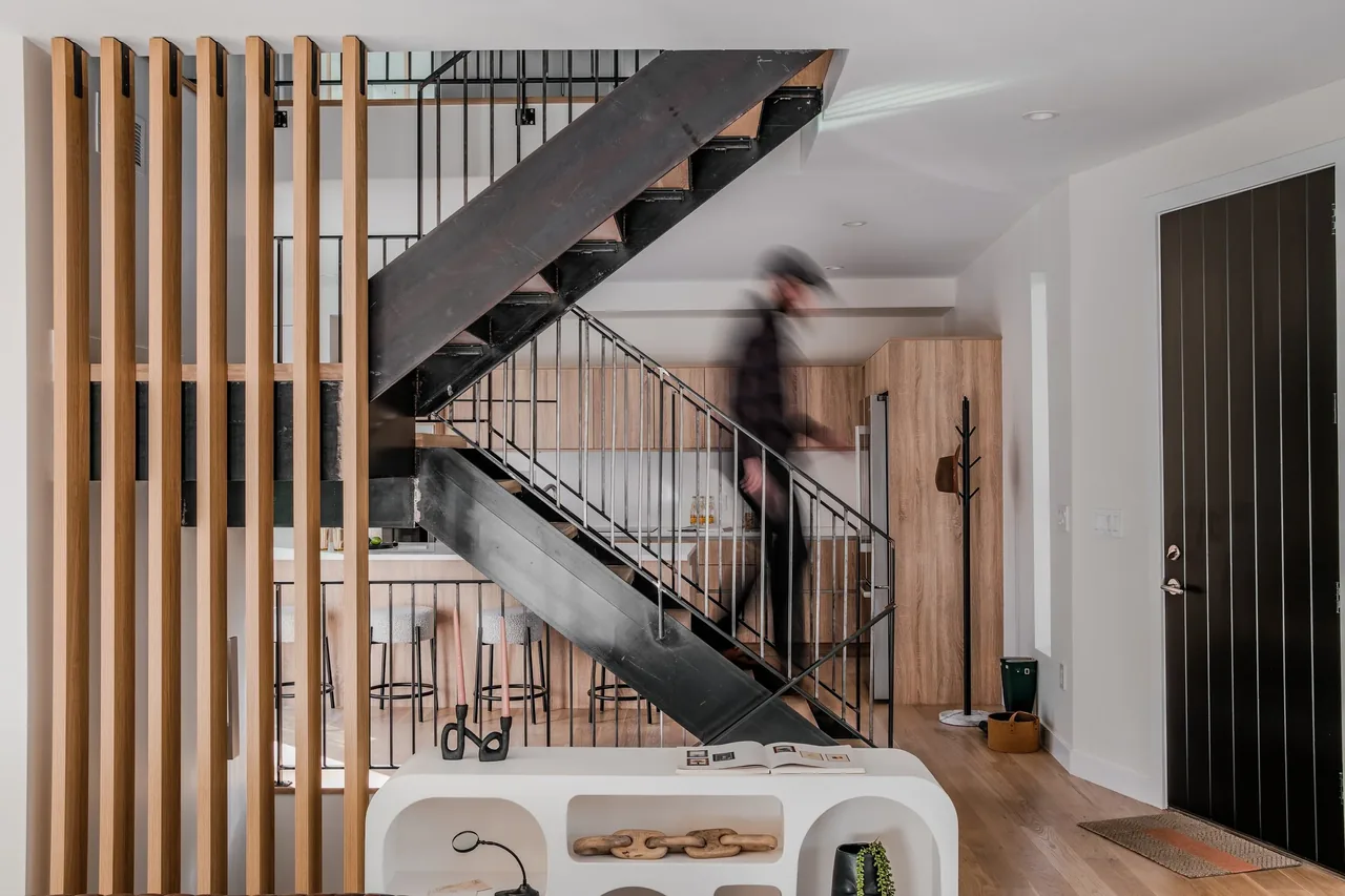 Modern interior with a black metal staircase, vertical wood slats, and a kitchen featuring wood cabinetry and bar stools.