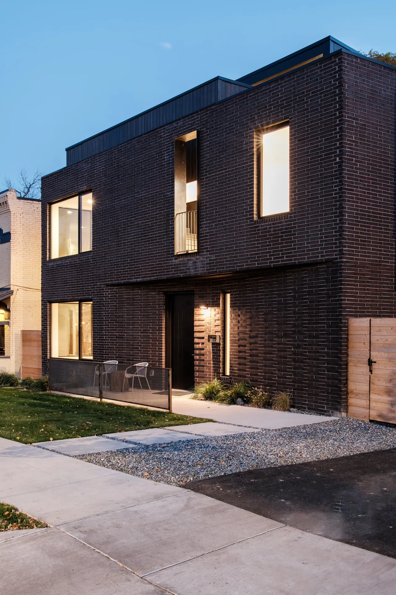 Two-story house with dark brick facade and vertical wood siding on upper volume, illuminated at dusk.