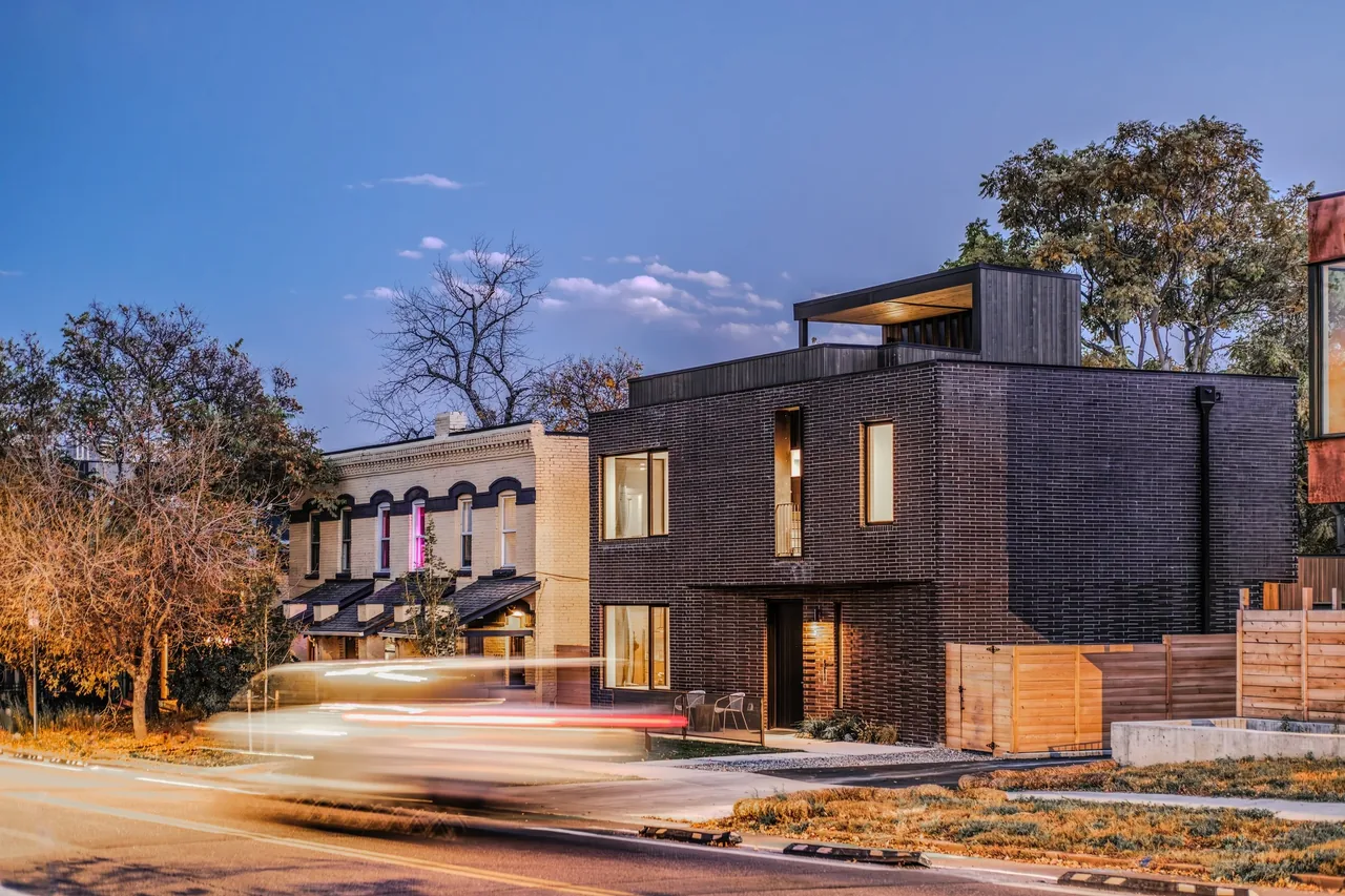 Modern residence featuring dark brick walls and a rooftop structure with wood siding next to older buildings.