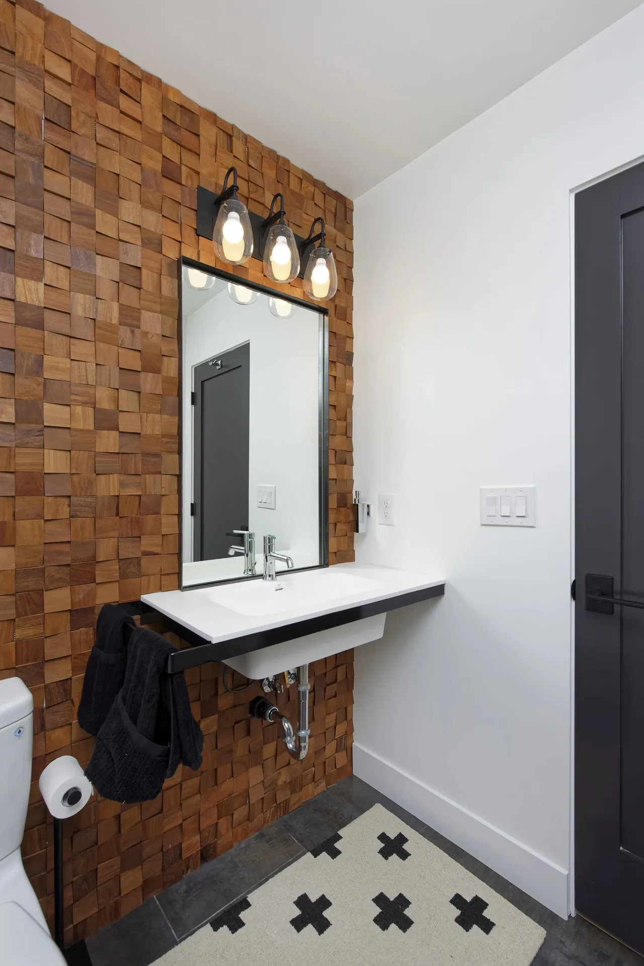 Bathroom features a wood textured accent wall with a rectangular mirror and triple light fixtures above the floating sink.