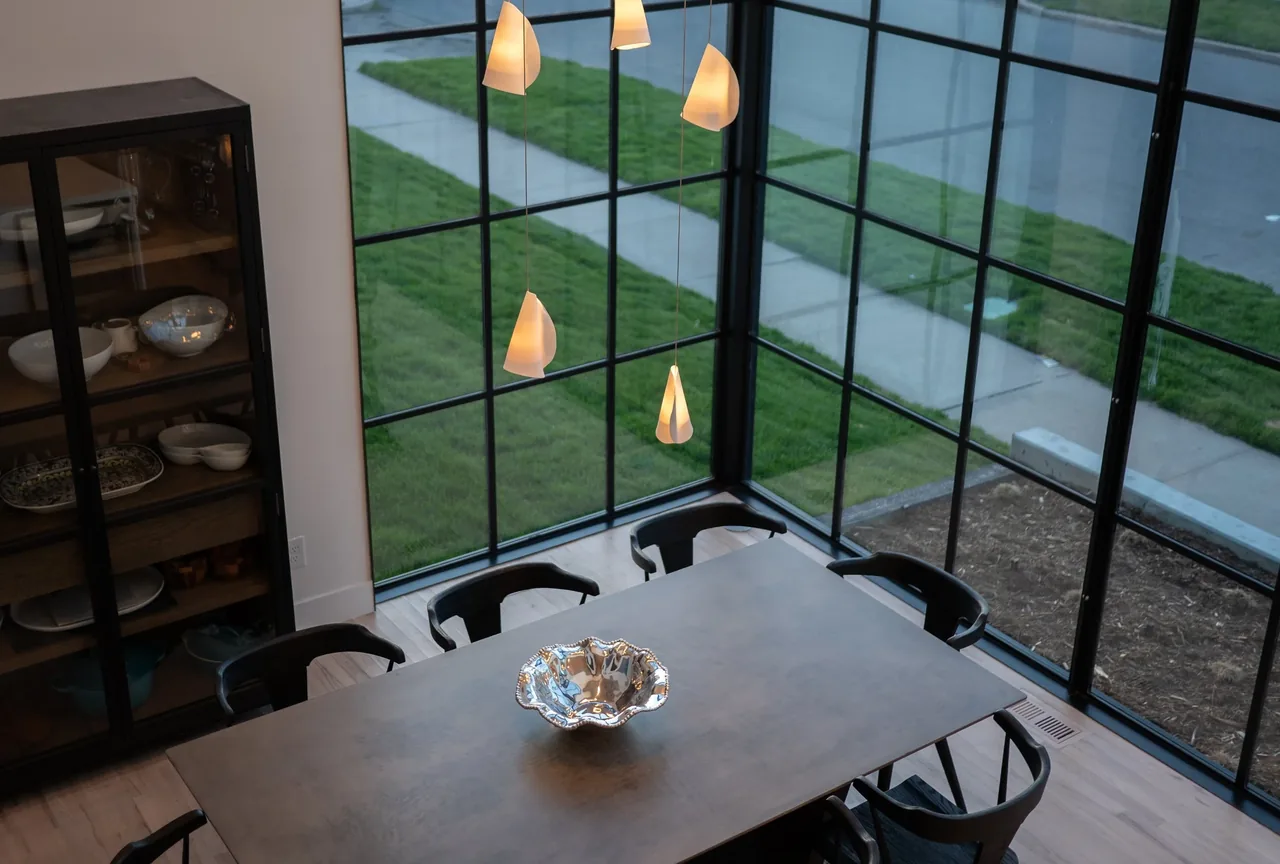 Dining area with a rectangular table and pendant lights, featuring floor-to-ceiling black-framed windows providing an exterior view.