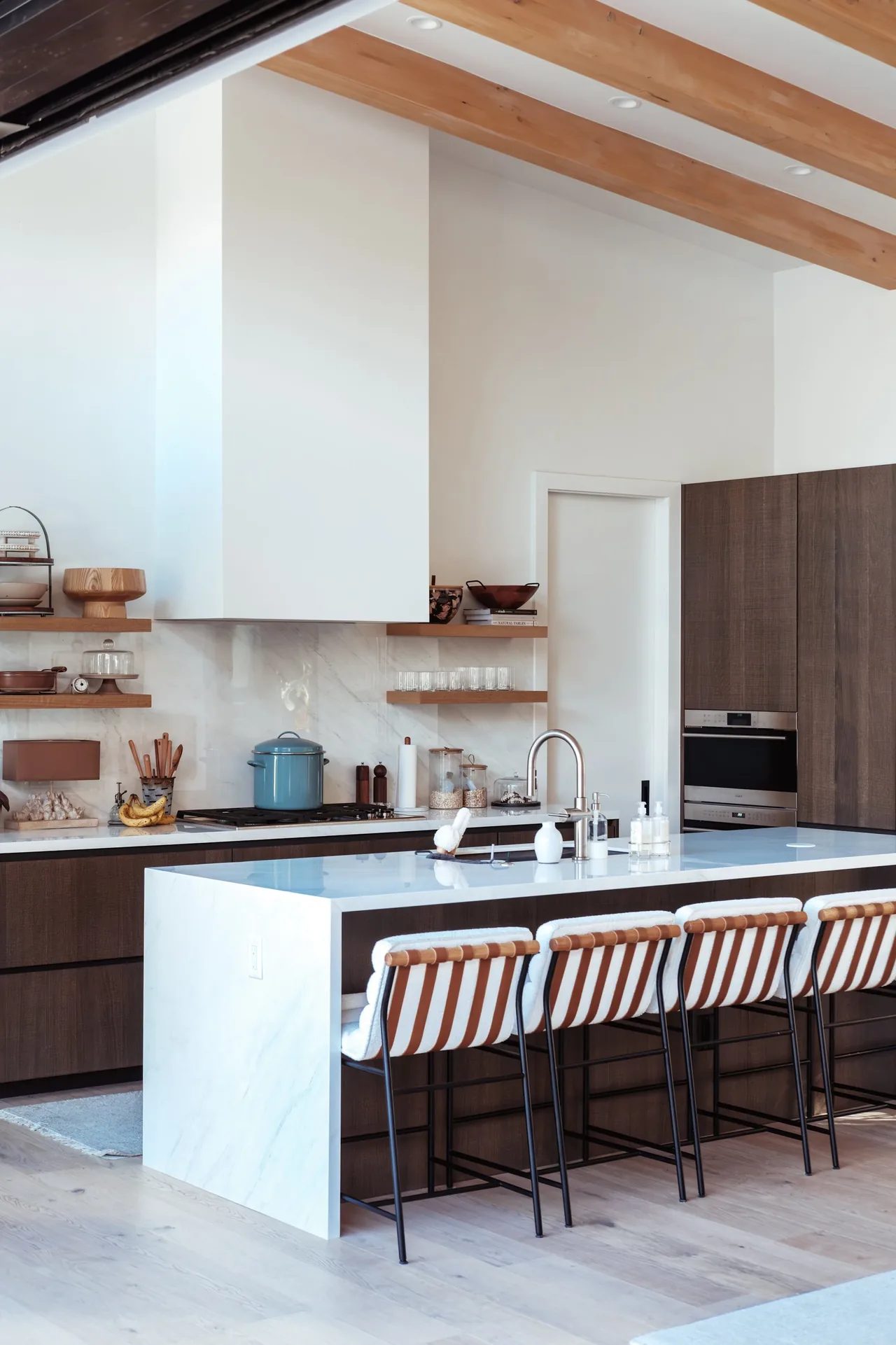 Kitchen featuring wood cabinets, a white marble island with a sink, wood ceiling beams, and open shelving.