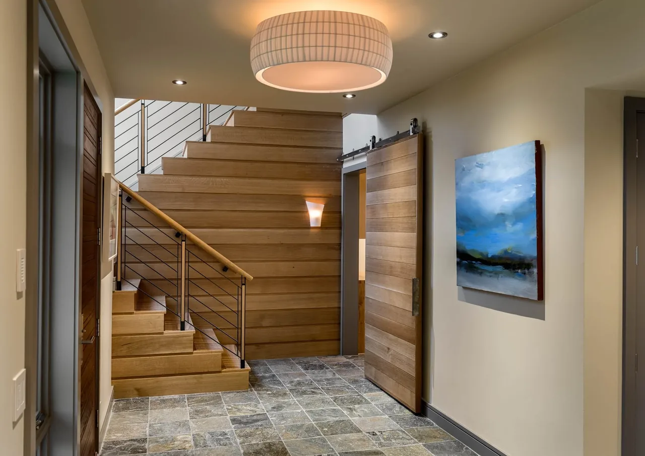 Entryway features wood stair with metal railing, a wood sliding barn door, stone tile floor, and wall art under ambient lighting.