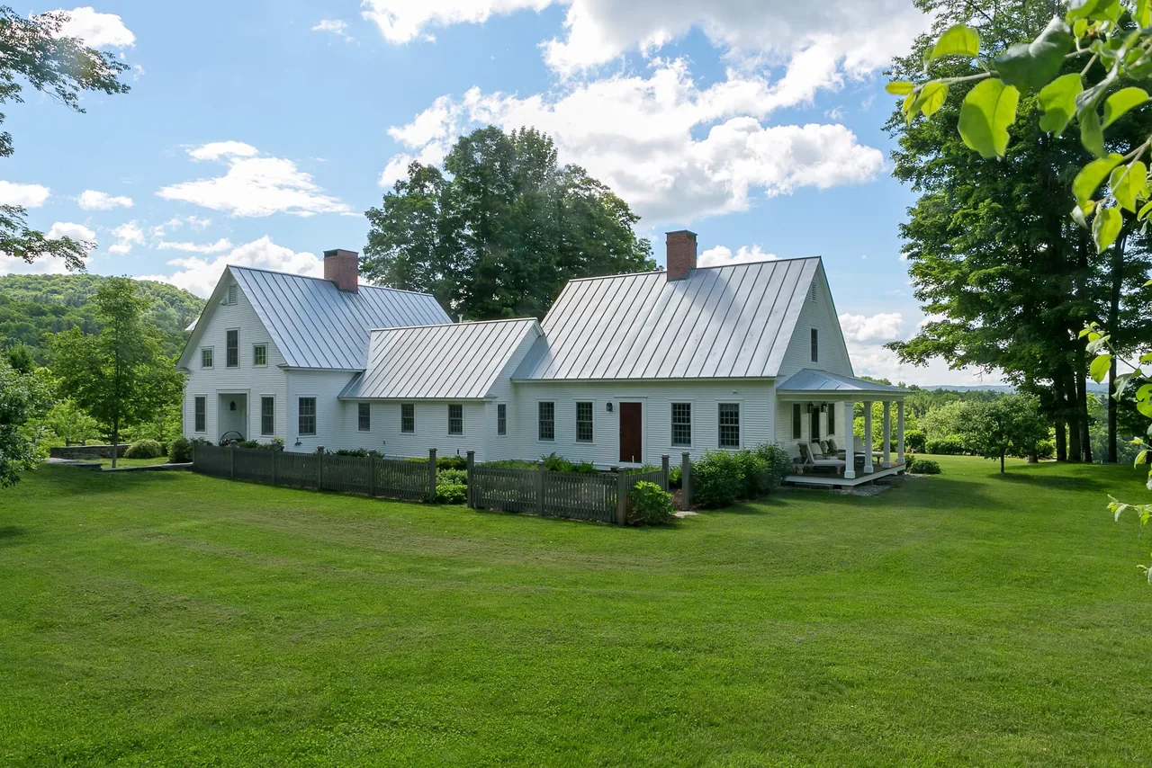 The Corinth Retreat features a traditional white farmhouse with standing seam metal roofs and a wood fence enclosing a garden area.