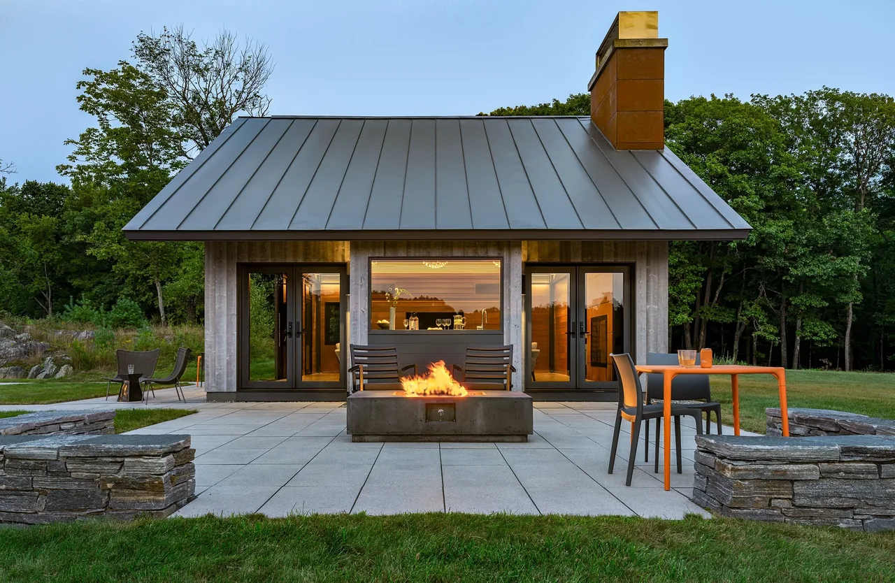 A small modern building with a standing seam metal roof and metal-clad chimney, featuring an outdoor seating area with a fire pit on the patio.