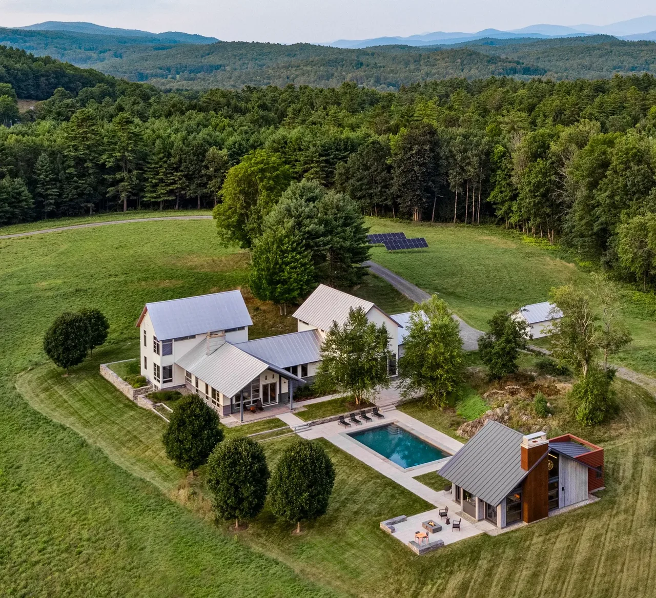 Lake retreat with multiple gable roof structures, an outdoor pool, and solar panels, set in a wooded landscape.