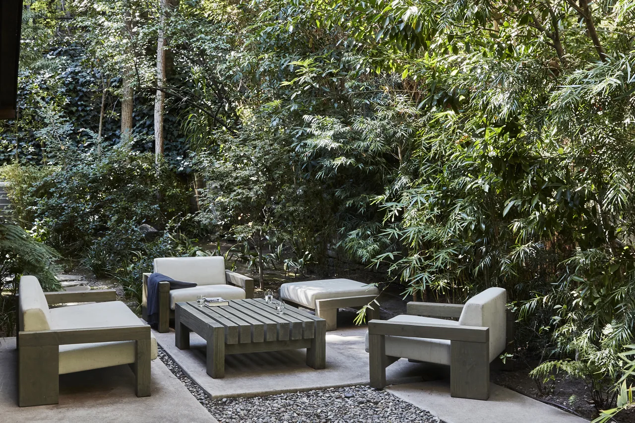 Outdoor living space featuring wooden framed furniture with cushions amid dense natural landscaping.