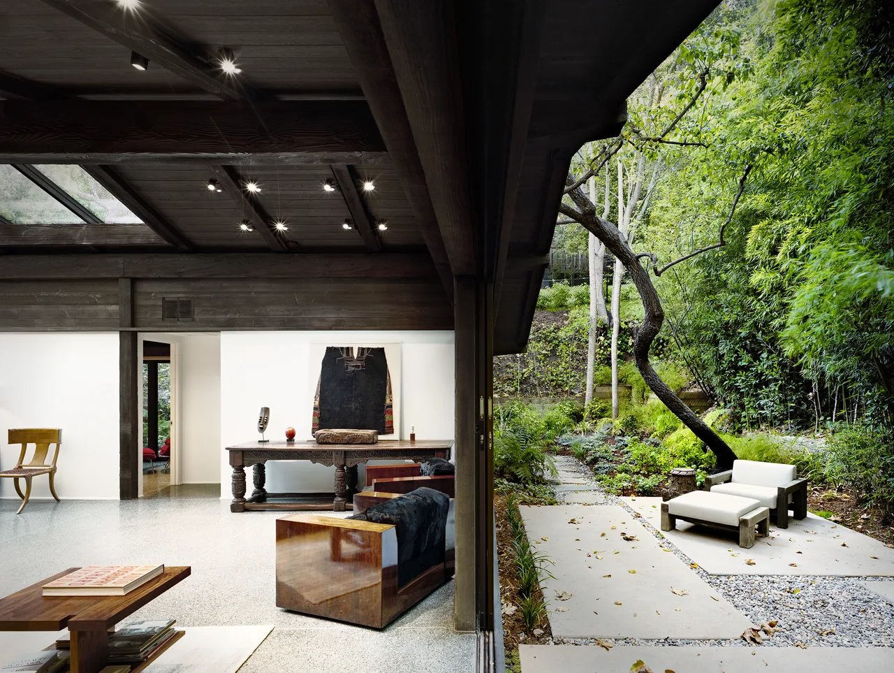 Living room with exposed dark wood beams opens to an outdoor patio surrounded by dense green landscaping.
