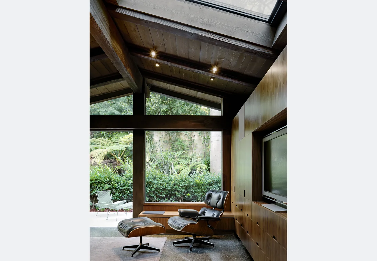 Living room features vaulted ceiling with exposed beams, floor-to-ceiling glazing with views to lush landscaping, and built-in wood cabinetry with a TV.