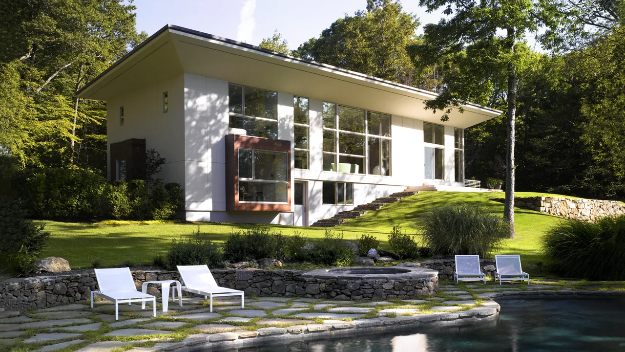 The Pawling Residence features a modern design with large floor-to-ceiling windows and a wide roof overhang, set in a wooded landscape.