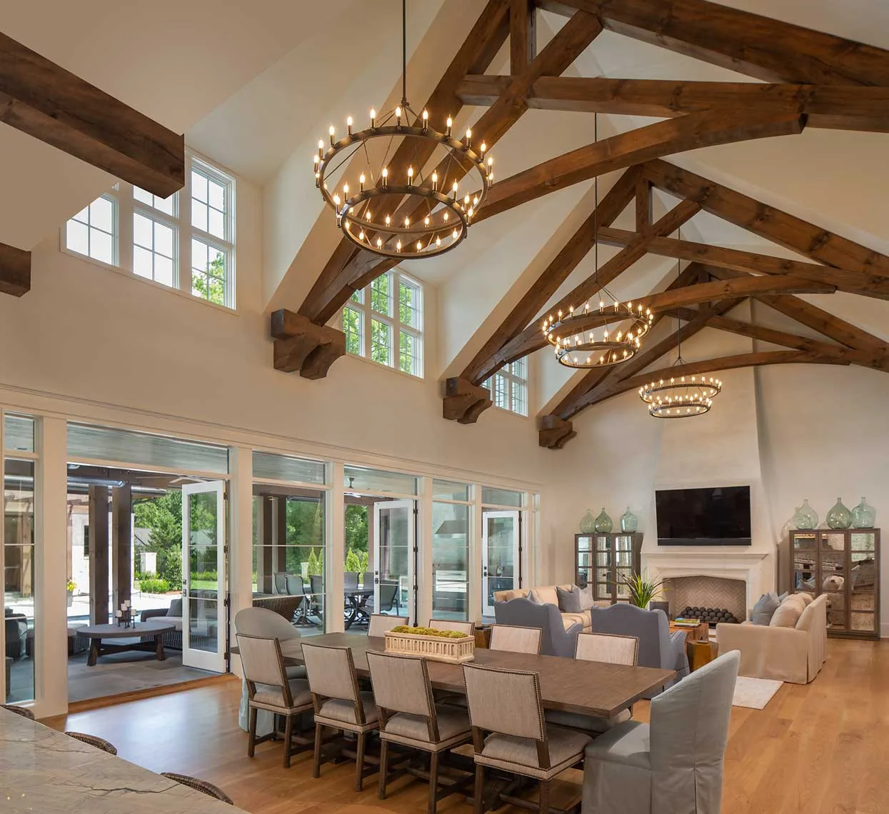 Open living and dining room with vaulted ceiling, exposed wood beams, circular chandeliers, and floor-to-ceiling windows overlooking outdoor seating.
