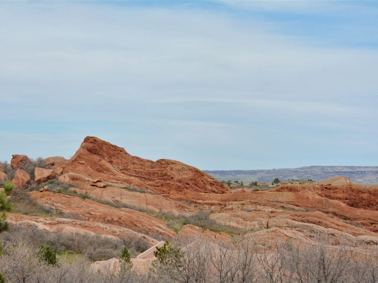 Natural red rock formations dominate the landscape with dry vegetation in front beneath a partly cloudy sky.
