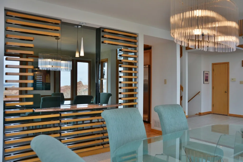 Dining area with a glass table, upholstered chairs, and a decorative wall mirror with horizontal wooden slats.