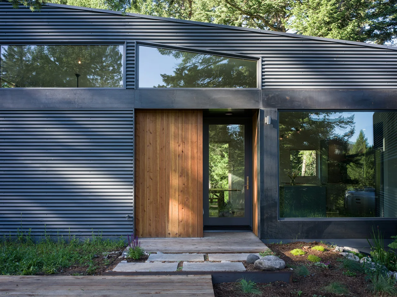 The Willow Creek Ranch House features corrugated metal siding with large windows and a wood front door set in a forest environment.
