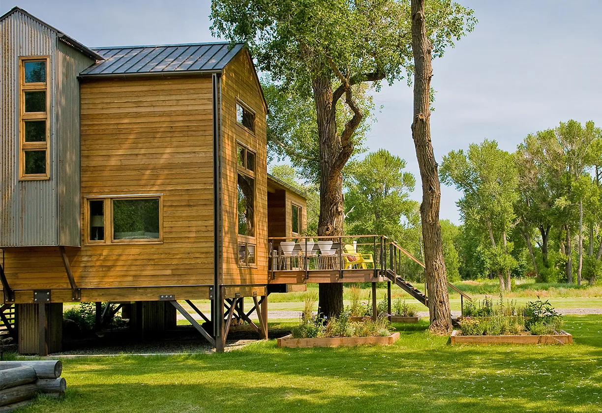 Yellowstone River Cabin features a stilt foundation and metal deck railing, set in a green landscape with mature trees.