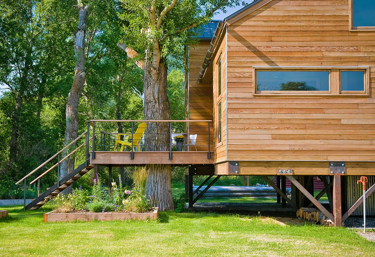 The Yellowstone River Cabin features a wood exterior with a deck on stilts and metal railing, blending into the natural landscape.