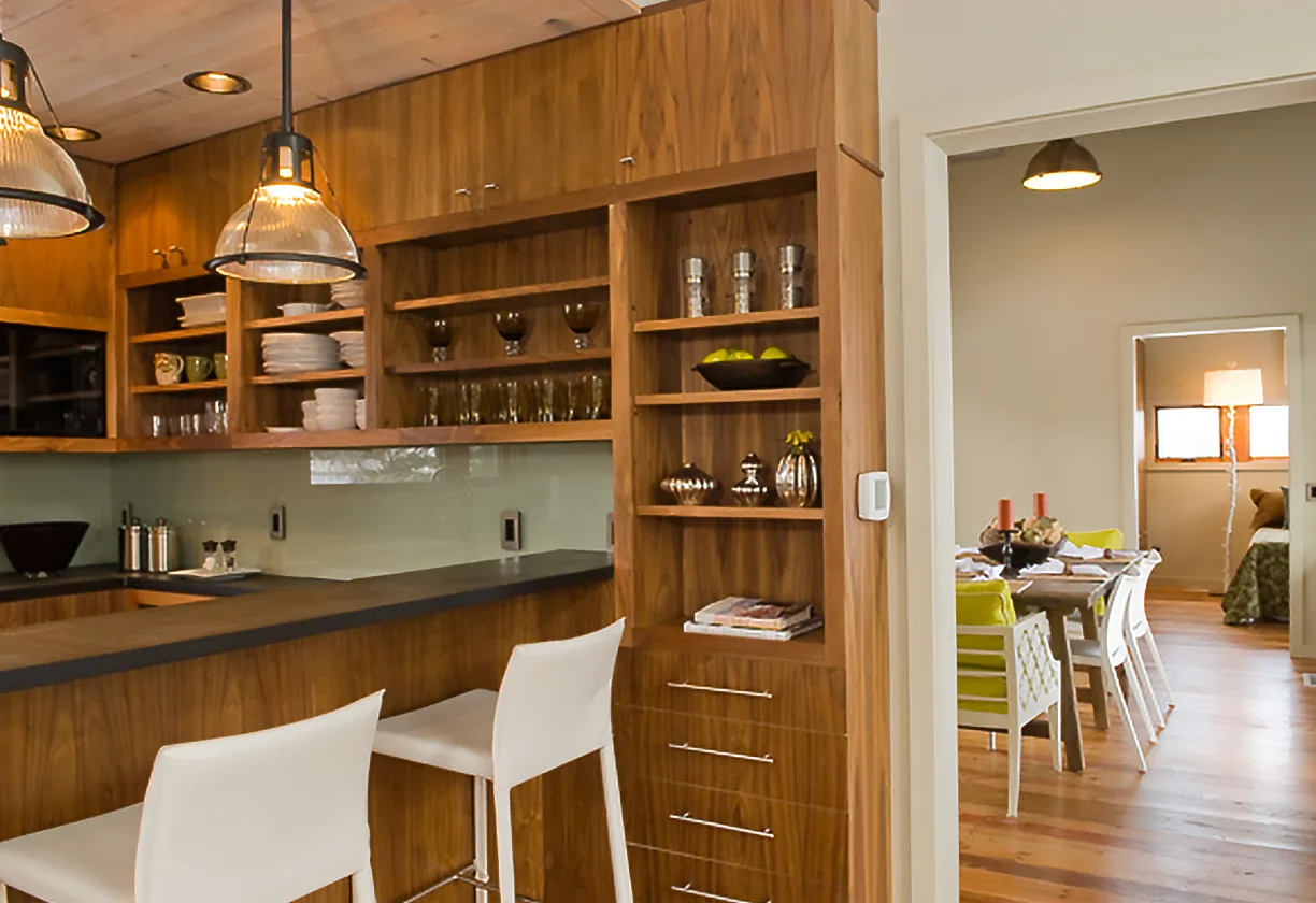 This image shows a wood-paneled kitchen with open shelving and barstools, adjacent to a dining room with green chairs and white walls.