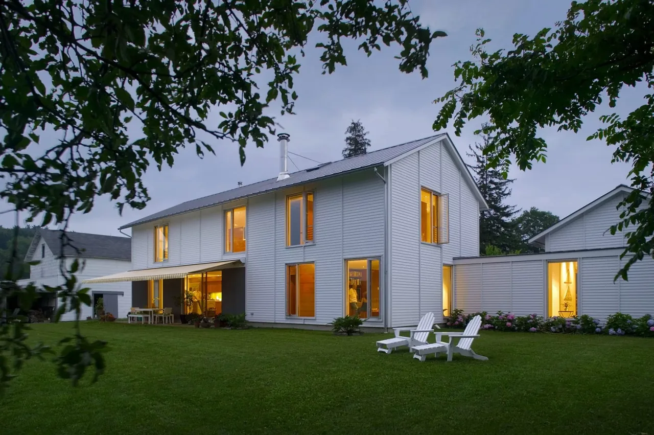 A two-story white house with a gable roof and outdoor seating on a grassy lawn in twilight.