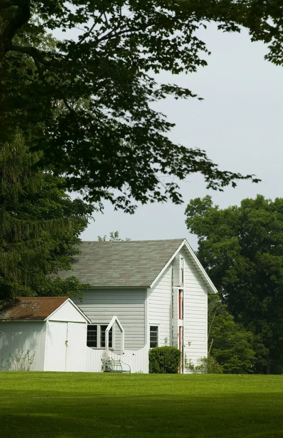 White wooden barn structure with adjacent shed and green lawn amidst large trees in a rural setting.