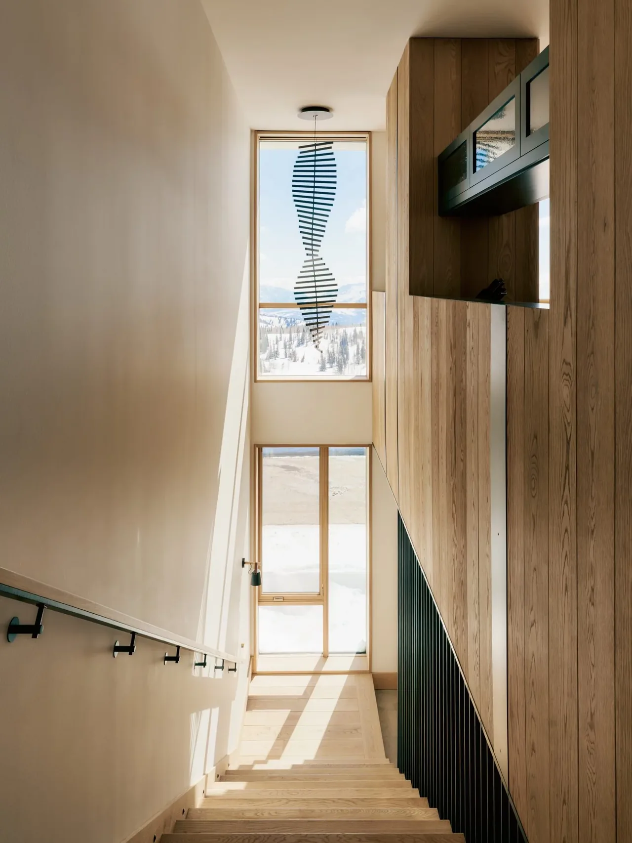 Natural light floods a wood-paneled stairwell with large vertical window facing snow-covered land.