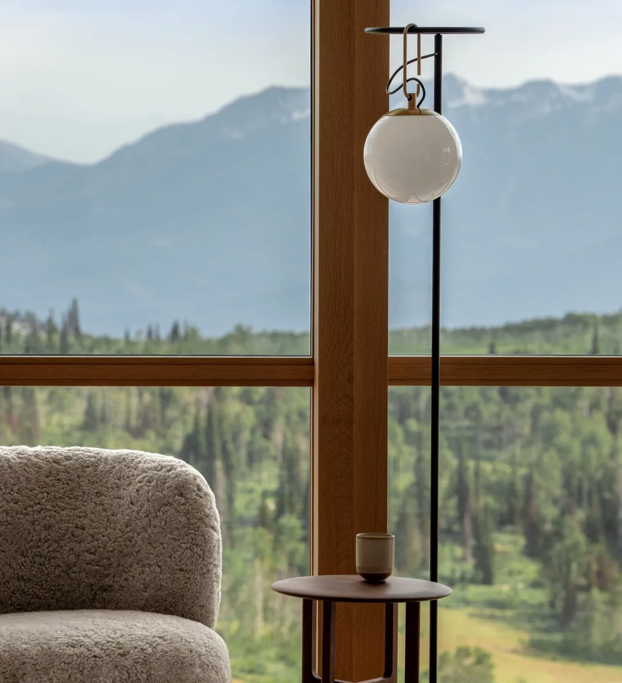 Interior detail featuring a textured armchair, round side table, and black lamp framed by a wooden window overlooking mountains.