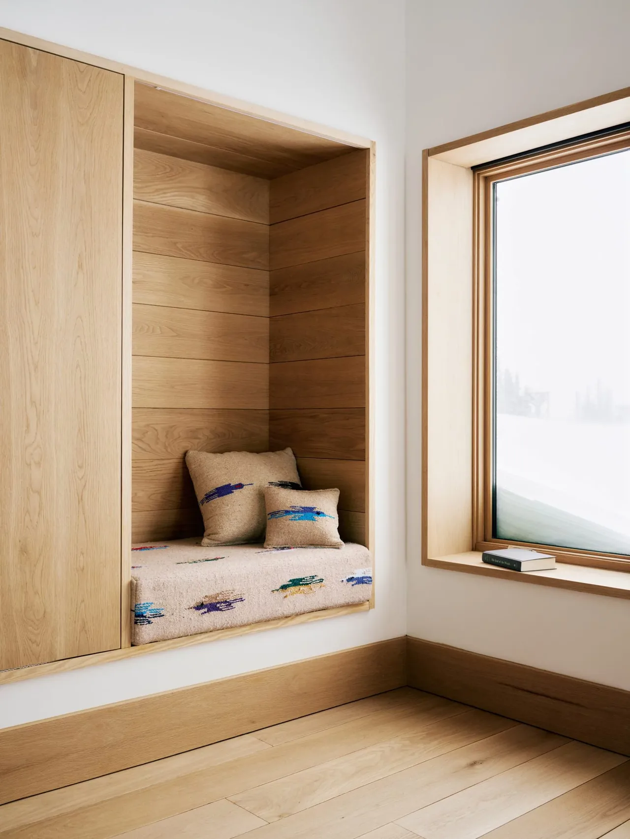 This living space features a built-in window seat with natural wood paneling and a large vertical window overlooking snow.