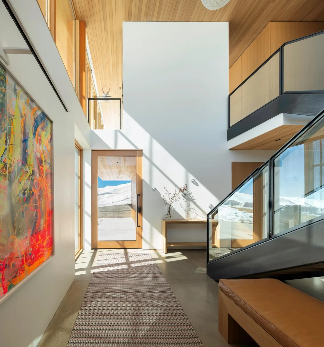 Entry hall with wood ceiling, large abstract artwork, glass railing on stairs, and a view of snowy hills through the wood-framed glass door.