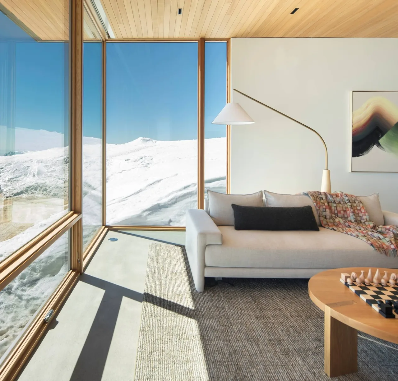 Living area with floor-to-ceiling wood-framed windows showing snowy hills outside, featuring a beige sofa and a curved white floor lamp.
