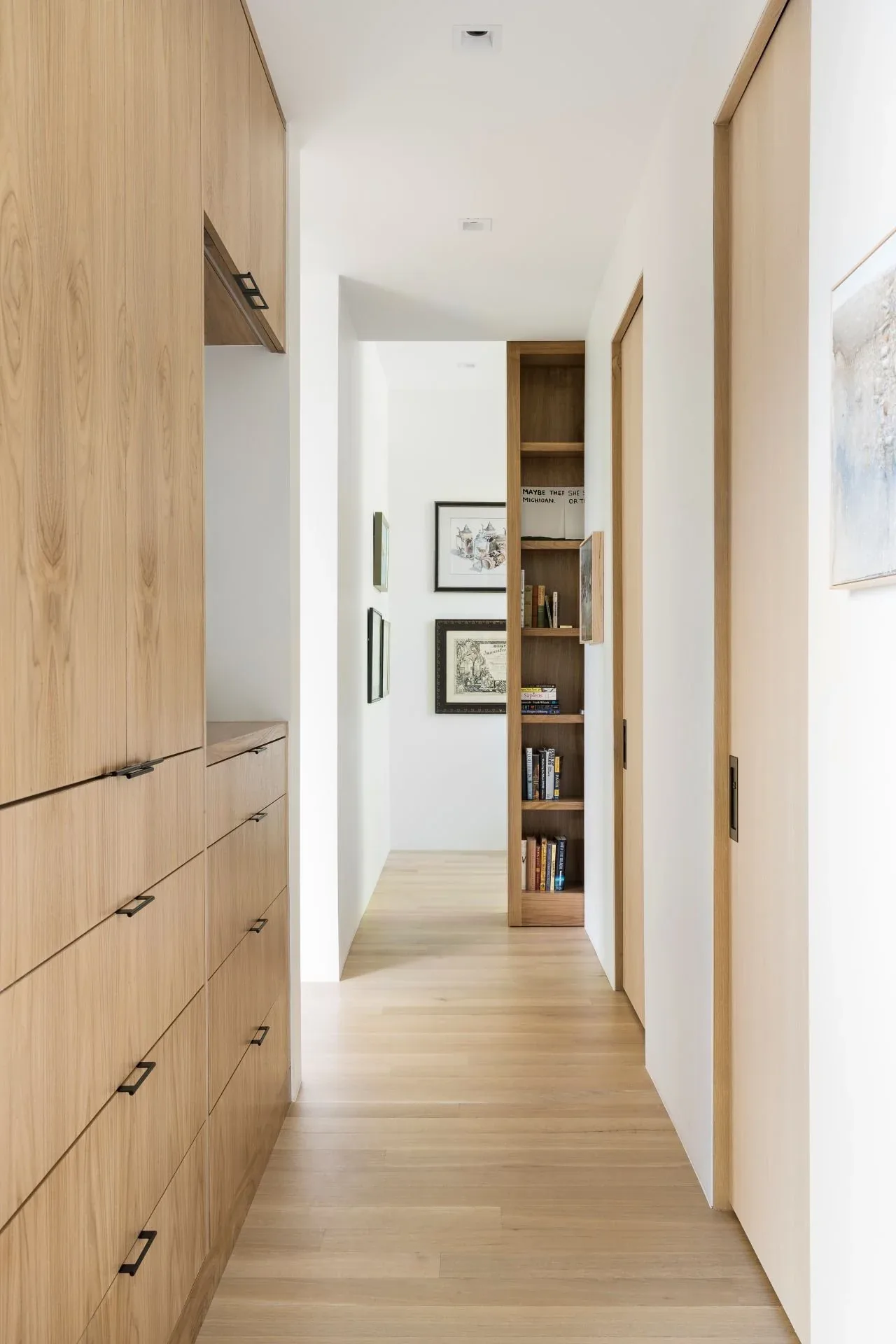 Hallway with light wood flooring, built-in wooden cabinets, and a recessed bookshelf with framed art on the walls.