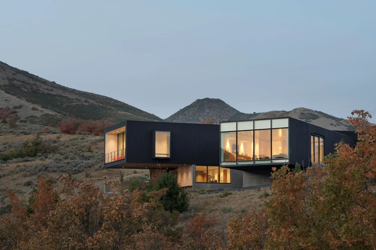 Hilltop house featuring black exterior with cantilevered design and large panoramic windows glowing at dusk.