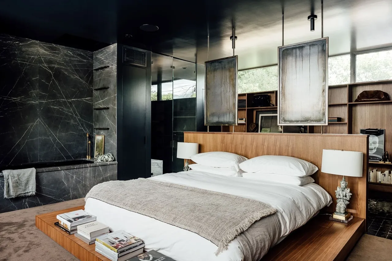 Bedroom with wood platform bed, black marble tub, built-in wood shelving, and contemporary artwork hanging from ceiling.