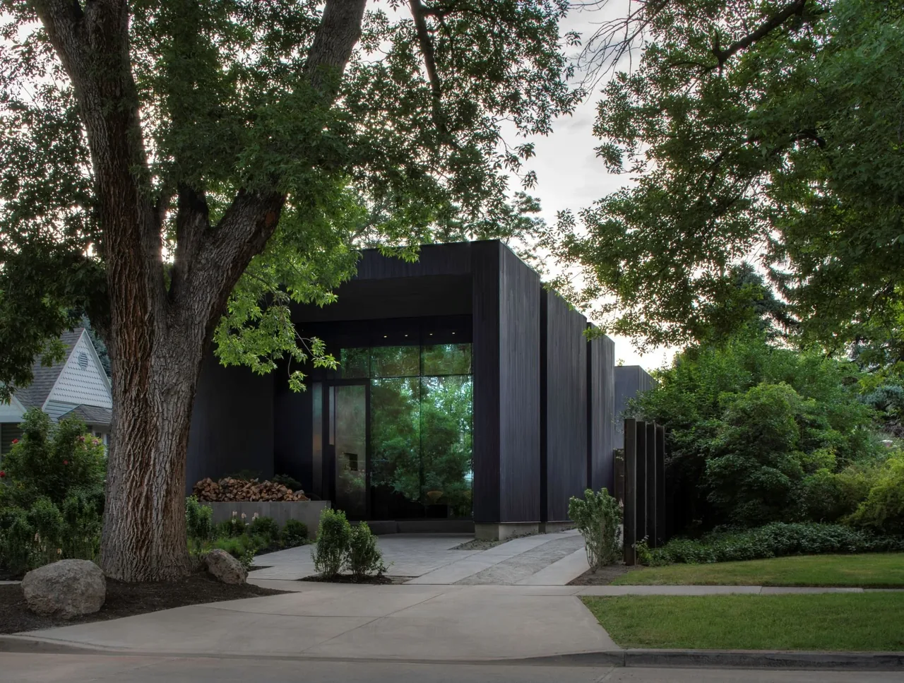 The Urban Treehouse features dark vertical siding and a large glass entry framed by mature trees.