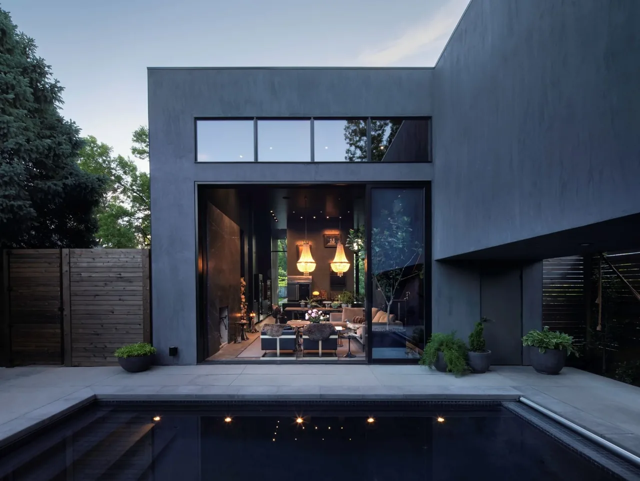 Modern home exterior showing black window frames, concrete steps, and a glass wall opening to the living room with pendant lights.