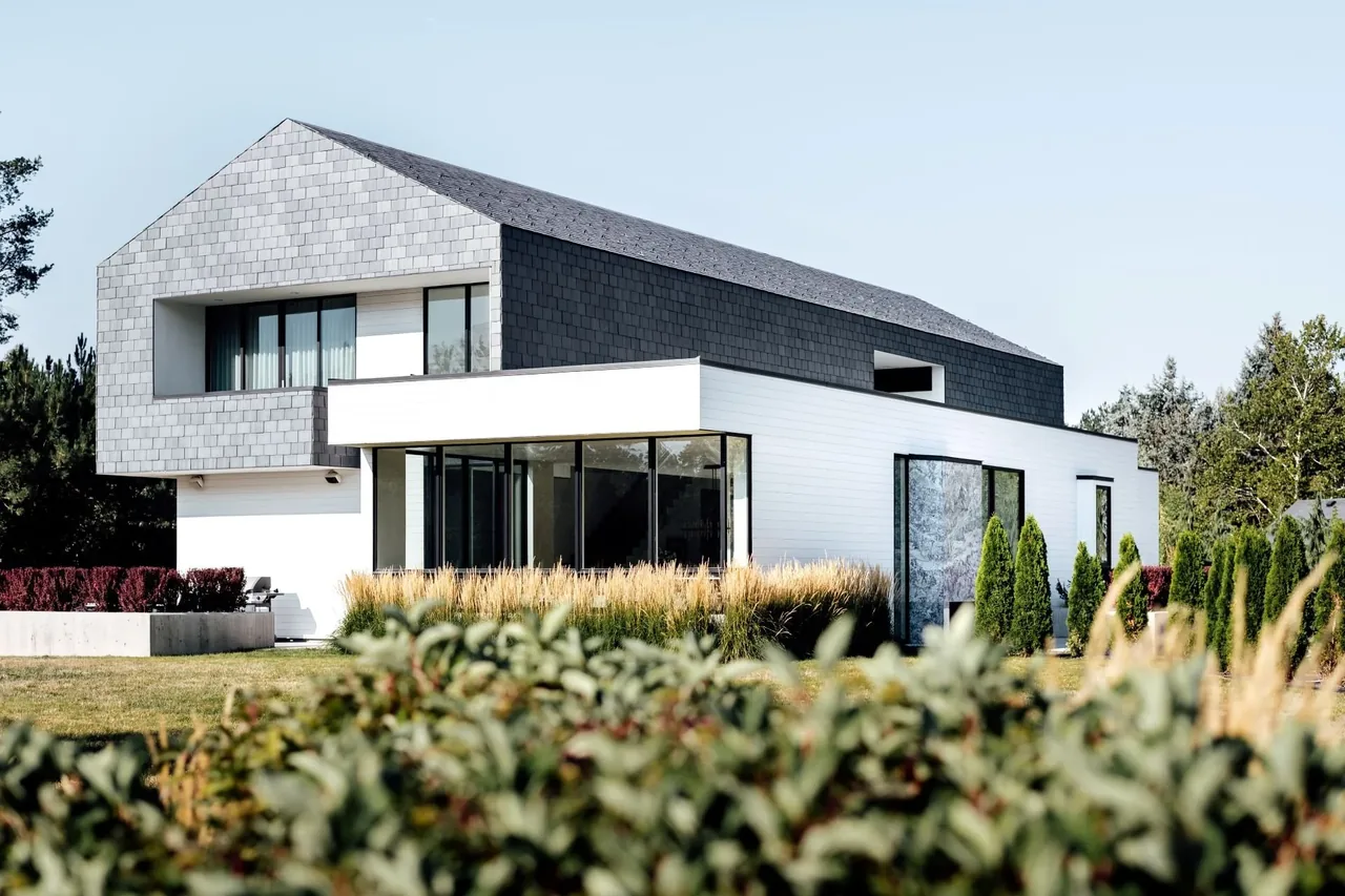 Modern farmhouse with dark slate shingles on the upper floor and white wood siding below, featuring large windows and landscaping bushes.