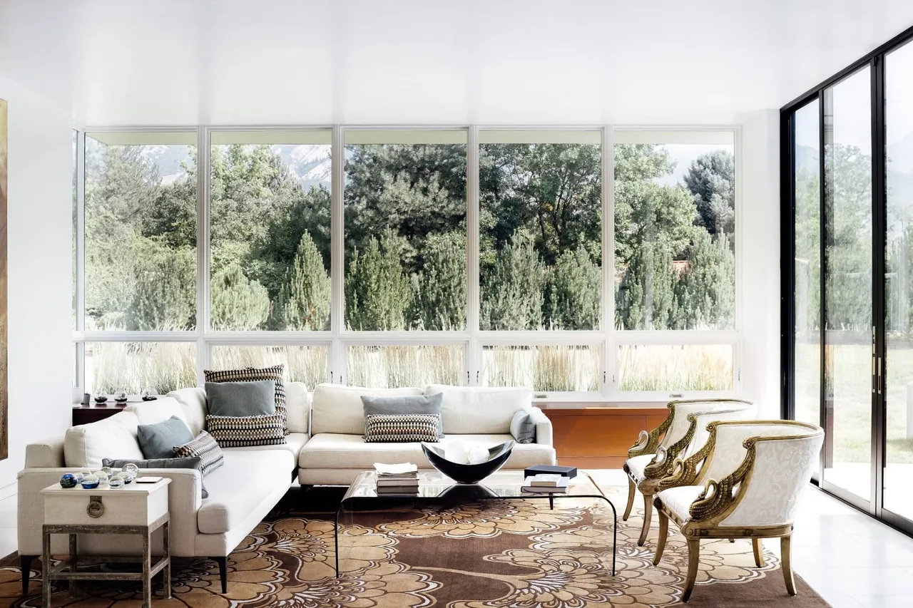 The modern farmhouse living room features expansive windows with views of lush greenery and a brown floral rug.