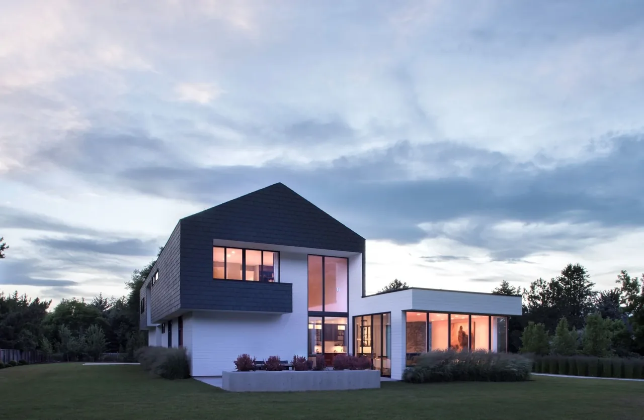 Modern farmhouse with black gable roof and white siding, featuring large windows and concrete planters with landscaping bushes.