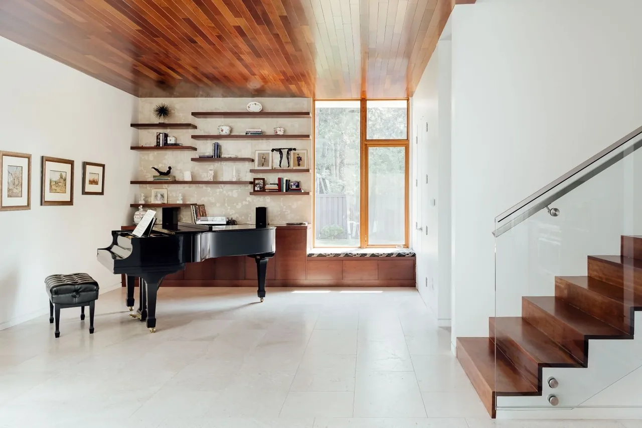 Living room features a black grand piano, wood paneled ceiling, floating shelves, and wooden stairs with glass railing.