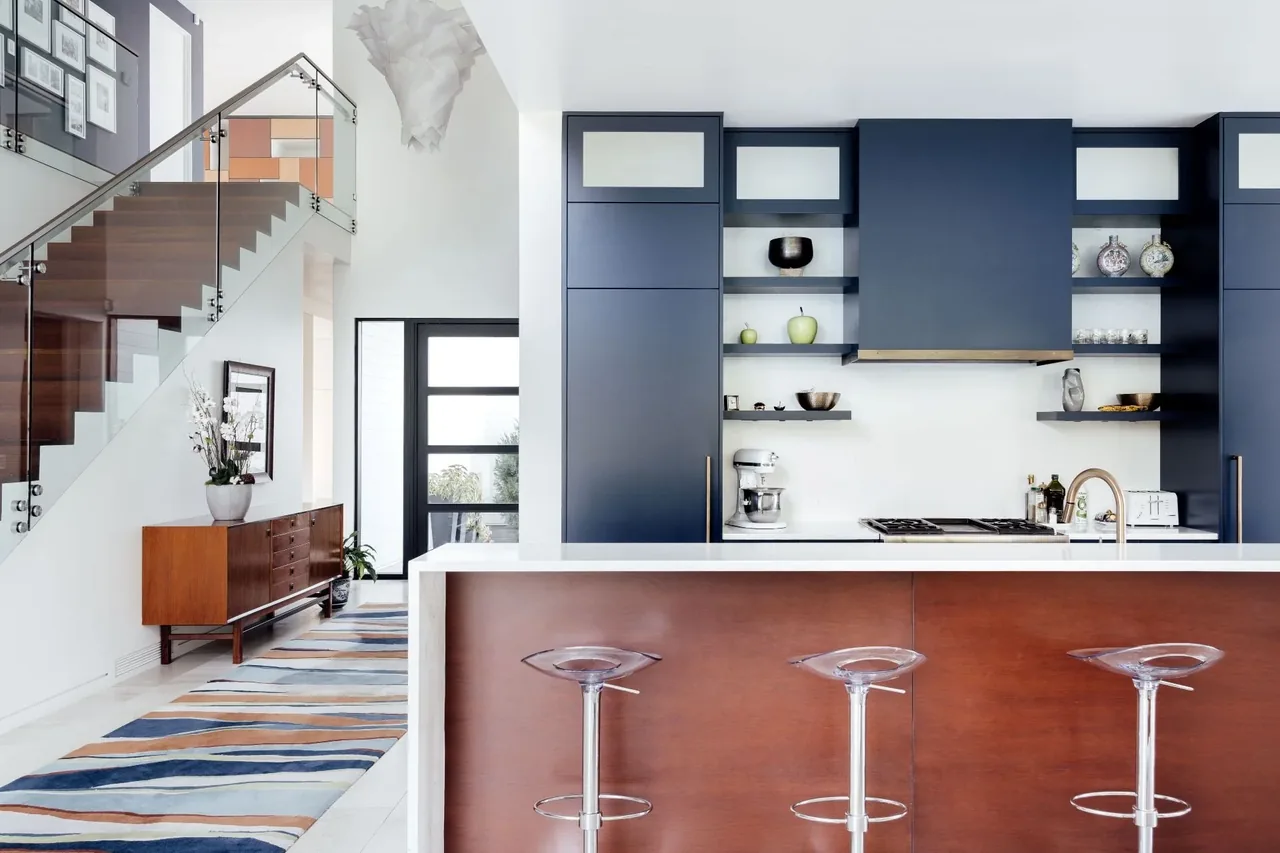 Kitchen features navy blue cabinetry with open shelving and a wooden island facing a glass-railed staircase.