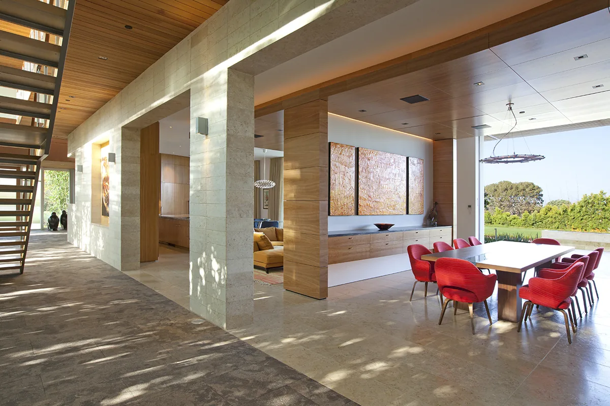 Dining area with red chairs and wooden ceiling opens to a landscaped lawn beside a hallway with stone flooring and wood ceiling.