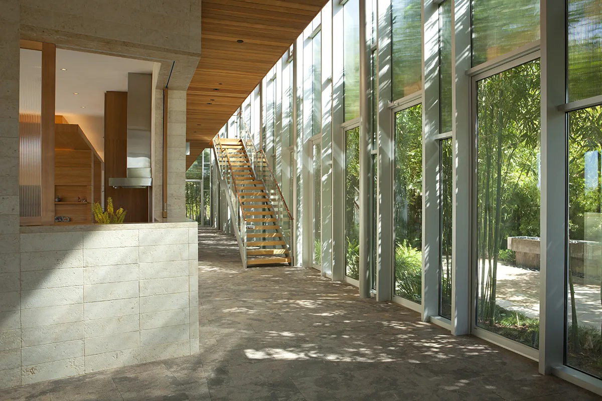 Long interior corridor features floor-to-ceiling glazing, a wooden staircase, and stone flooring beside a kitchen space.